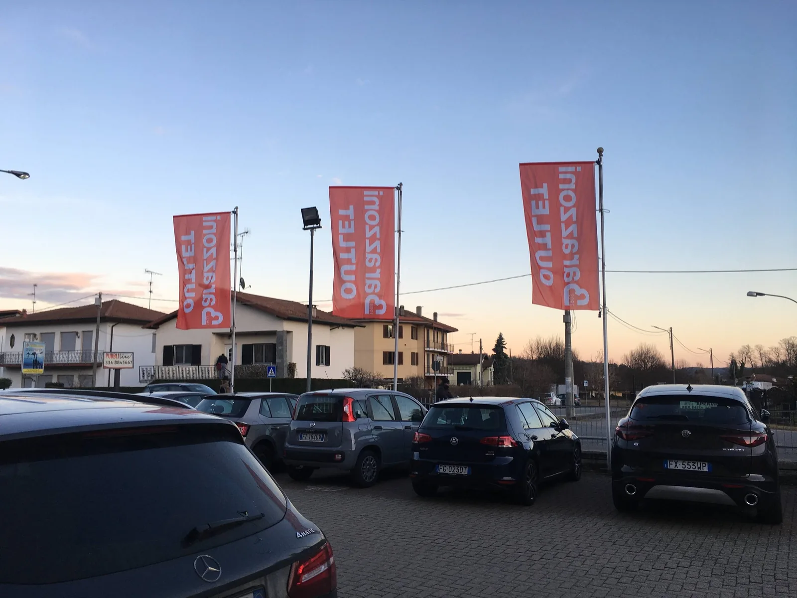 Tre bandiere verticali Barazzoni Outlet sui pennoni del parcheggio, al tramonto
