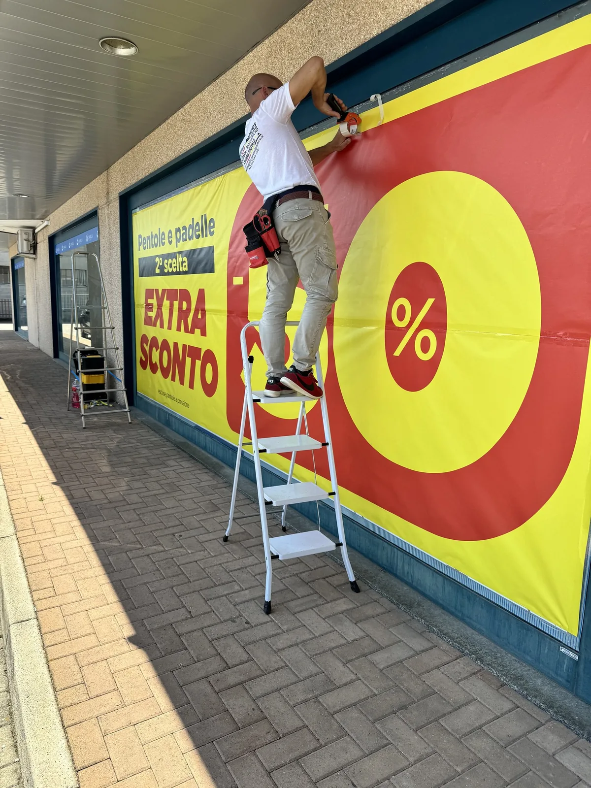 Posa del banner giallo e rosso Extra Sconto sulla vetrina, col posatore sulla scala