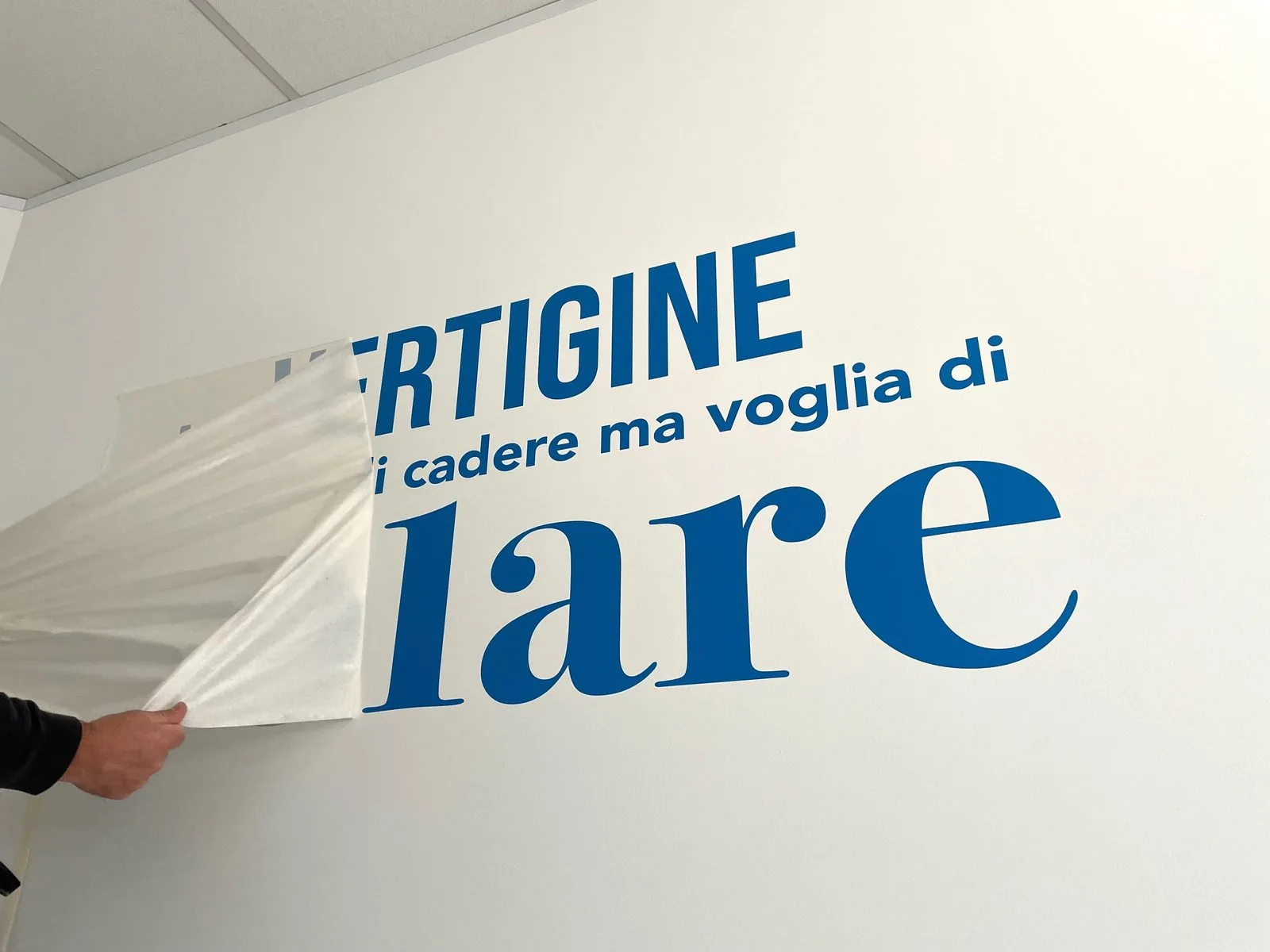 Dettaglio della scritta adesiva blu «Vertigine» mentre si stacca il supporto di trasferimento